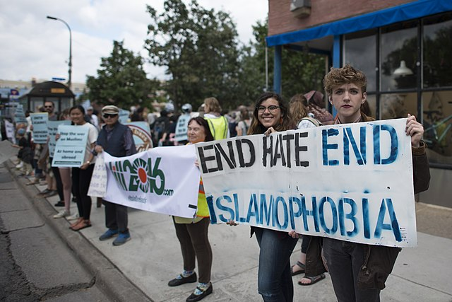 A group of diverse individuals stands on a sidewalk holding signs that read "End Hate, End Islamophobia."