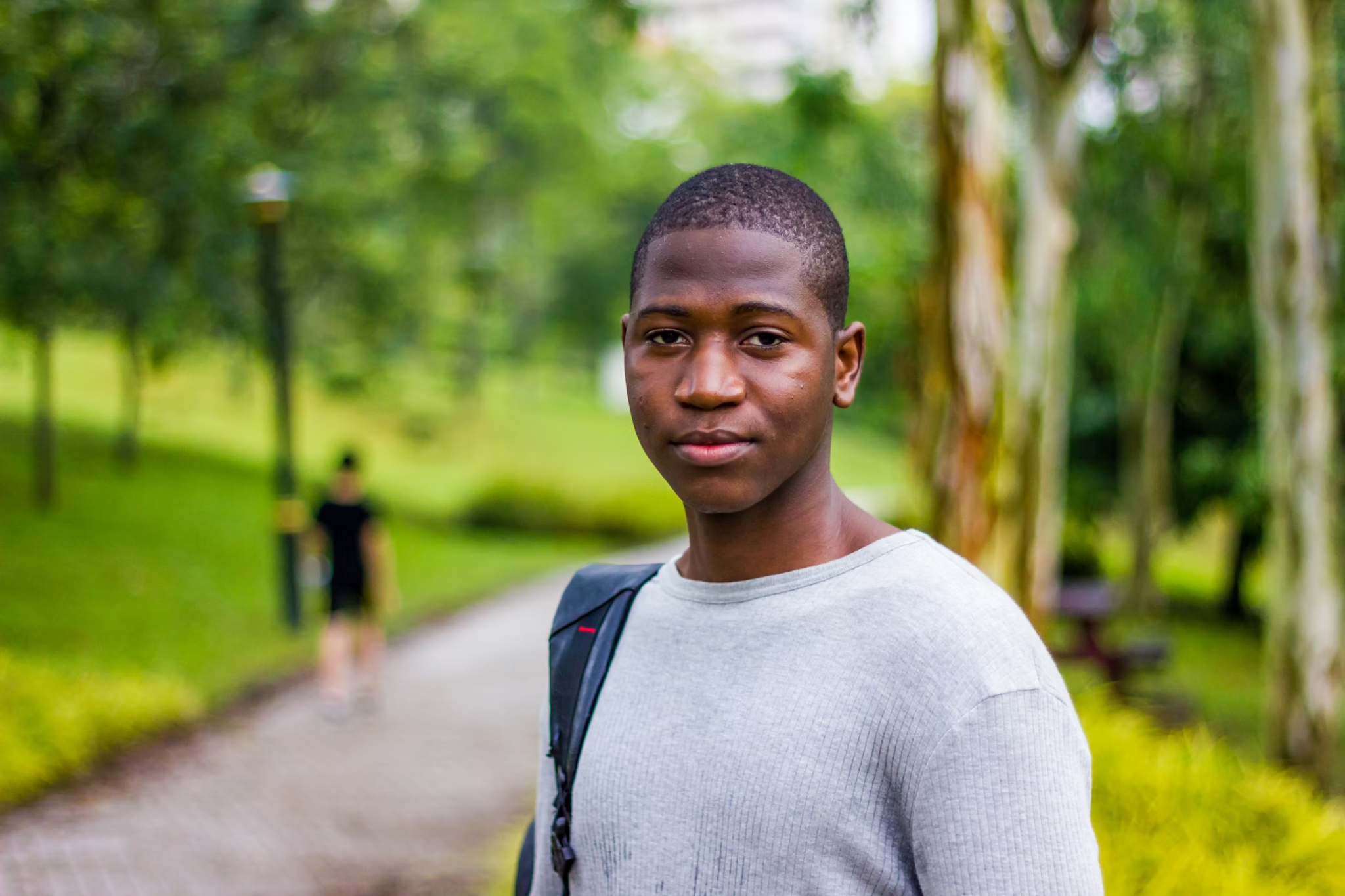 A young Black man stands outdoors on a park path, wearing a light gray sweater with a backpack strap visible.