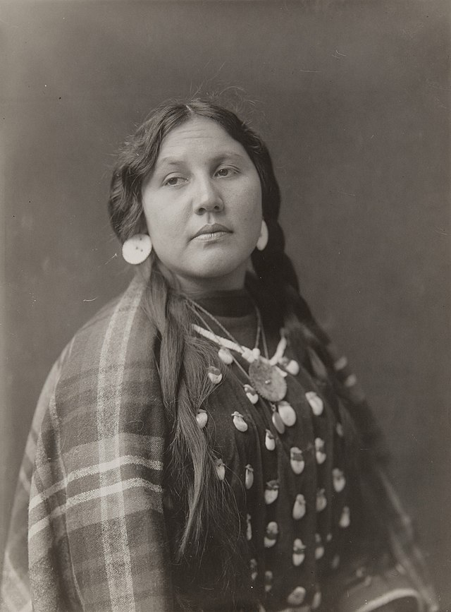 A sepia photograph of a Native American woman with long braided hair. She wears large earrings and a necklace, wrapped in a plaid shawl covered with shells. Her expression is calm and pensive.