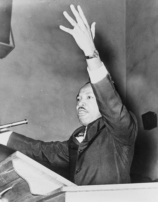 Martin Luther King Jr in a suit, half-length portrait, facing left, with left arm raised while speaking at a podium.