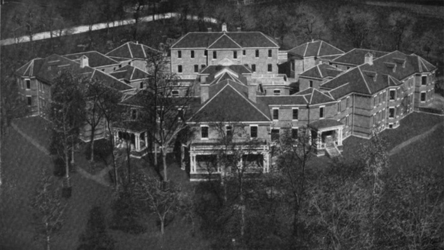 Aerial view of a large, historical building with multiple wings arranged in a circular pattern, surrounded by trees.