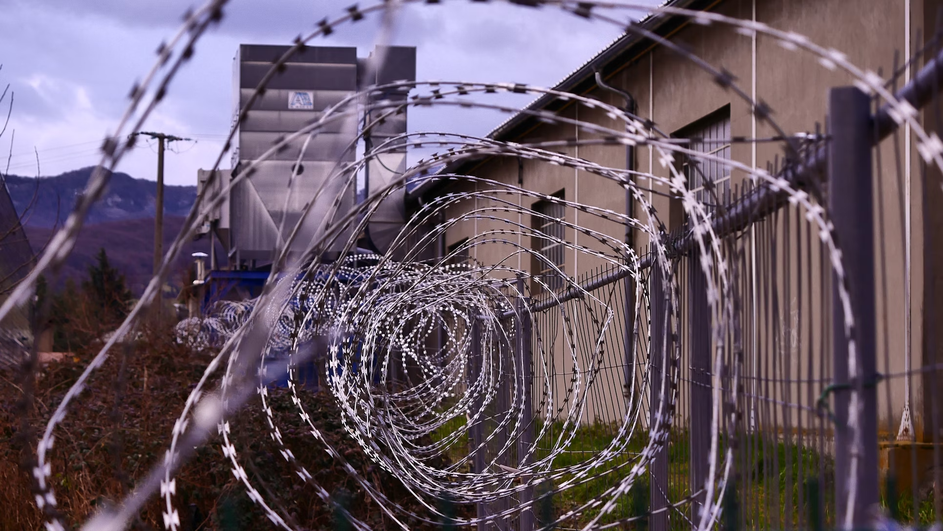 Spiraled razor wire atop a high fence surrounds an industrial building. The scene conveys security and confinement with a gloomy, overcast sky.