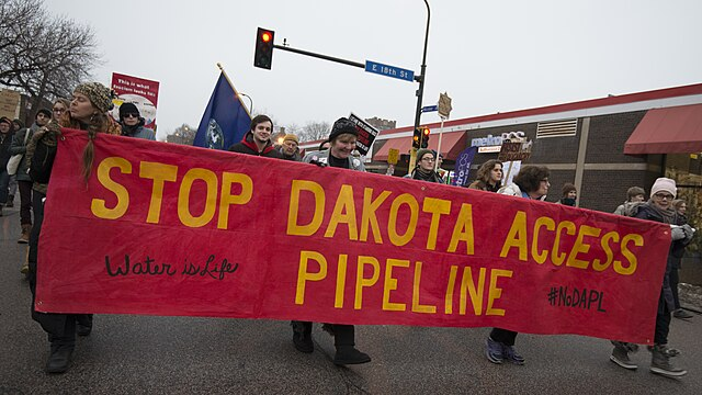A diverse group of protesters march down a street holding a large red banner reading "Stop Dakota Access Pipeline," and "Water is Life"