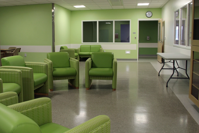 A bright, clean waiting room with light green walls and matching green cushioned chairs. The room feels calm with a clock on the wall and a white folding table.