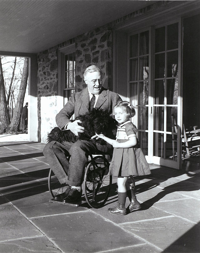 FDR shown in his wheelchair joyfully holds a black dog beside a young smiling girl on a sunlit porch. Stone wall and glass doors in the background.