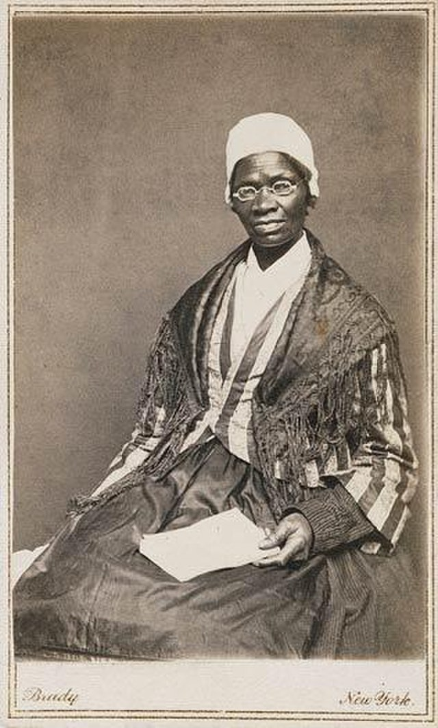 Sojourner Truth shown in an albumen silver print, wearing a shawl and bonnet, sits holding a book. Her expression is calm and composed and she looks directly at the camera.