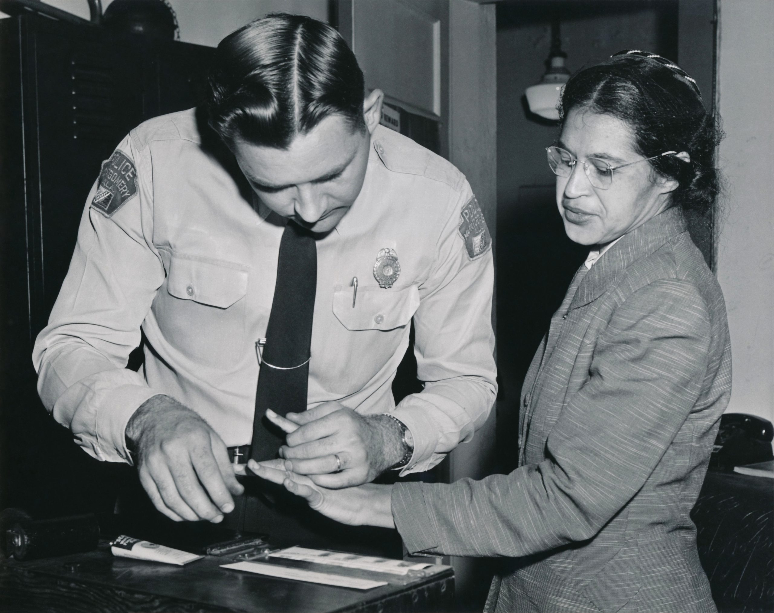 Black and white photo: a woman in a suit is having her fingerprint taken by a police officer.