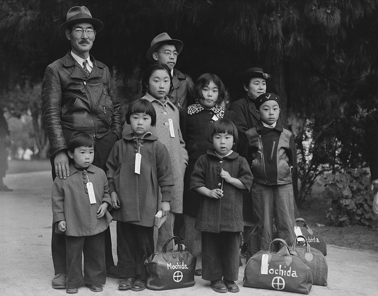A solemn Japanese family, with children and adults wearing coats and ID tags, stands beside luggage labeled "Mochida."