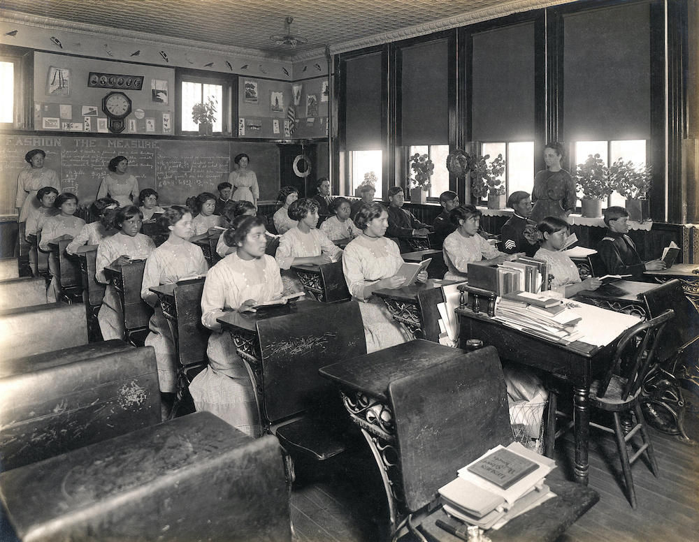 Historic classroom scene with Native American students in rows of wooden desks. Female teachers stand by a chalkboard.