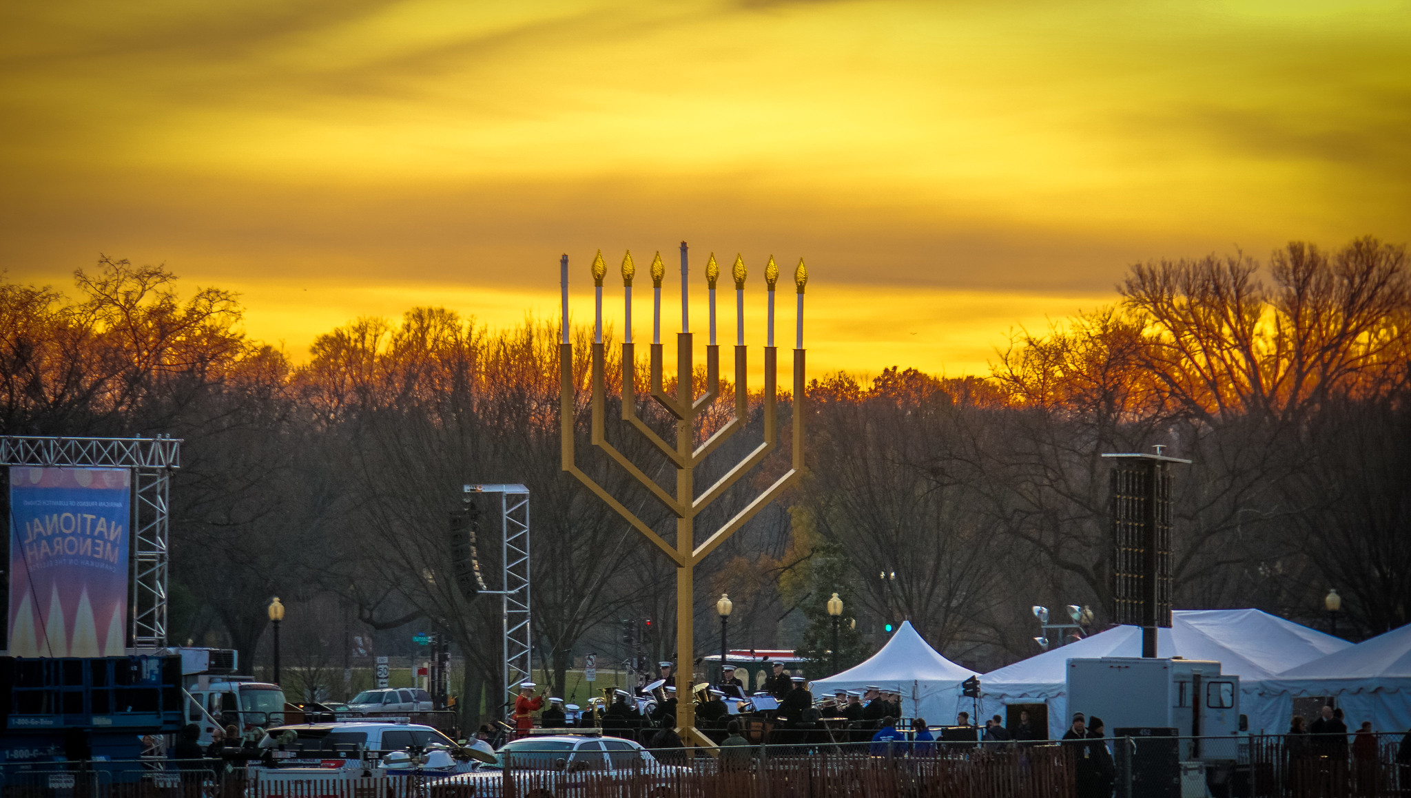 A large menorah stands prominently in front of leafless trees at sunset. The sky glows orange, creating a warm, festive atmosphere. Tents and people surround the menorah