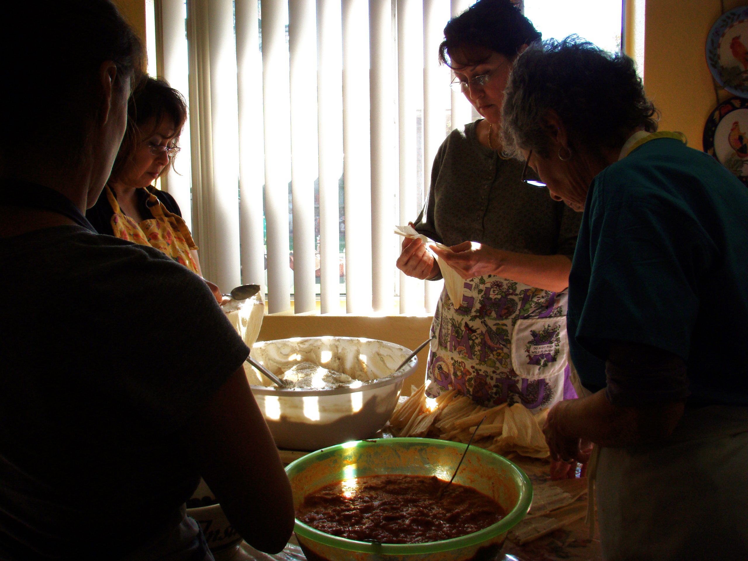 Four individuals gather around a table, preparing food in a softly lit room. They are focused on making tamales with aprons and ingredients spread out.