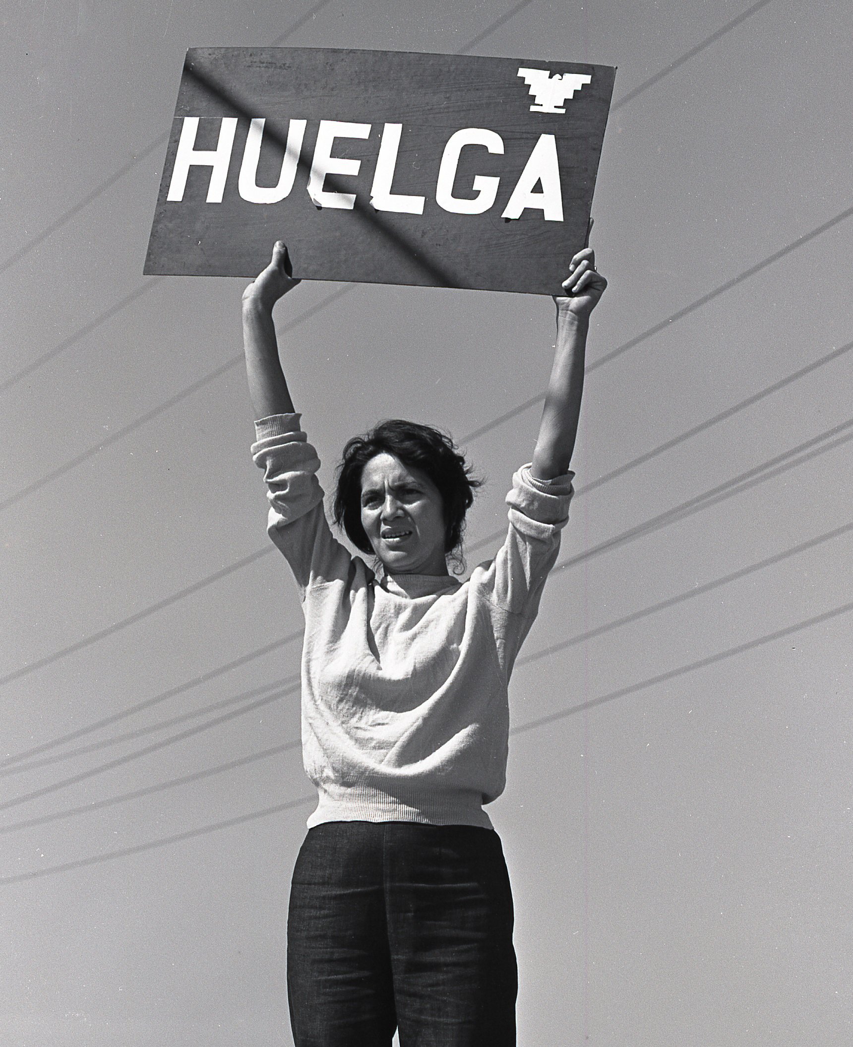 A person holds a sign reading "HUELGA" high above their head, standing against a clear sky.